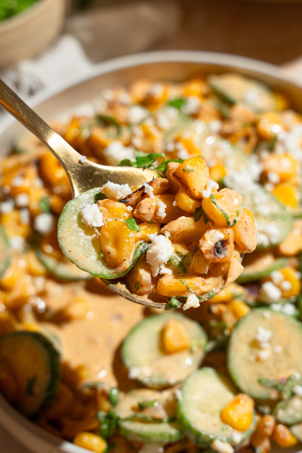 A close up of a spoon with a bite of Mexican cucumber salad. There is a bowl of salad tossed with seasoned corn, red onion, and cilantro in an elote dressing blurred in the background.