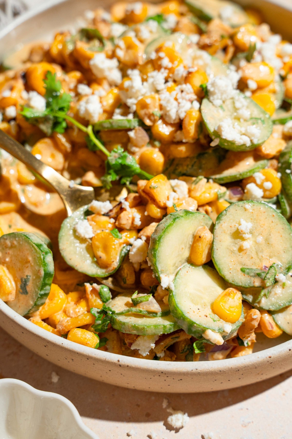 A white bowl with Mexican cucumber salad tossed with corn, cilantro, and onion tossed in an elote dressing and topped with cotija cheese. The bowl is on a tan counter.