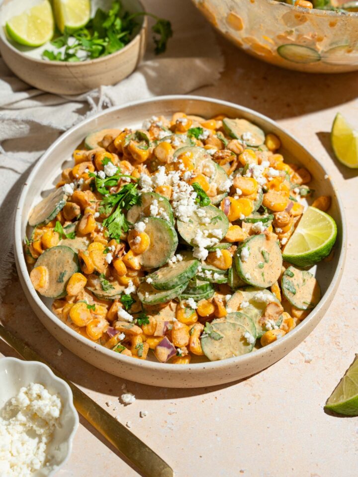 A white bowl with Mexican cucumber salad tossed with corn, cilantro, and onion tossed in an elote dressing and topped with cotija cheese. The bowl is on a tan counter with a bowl of cheese and bowl of toppings.