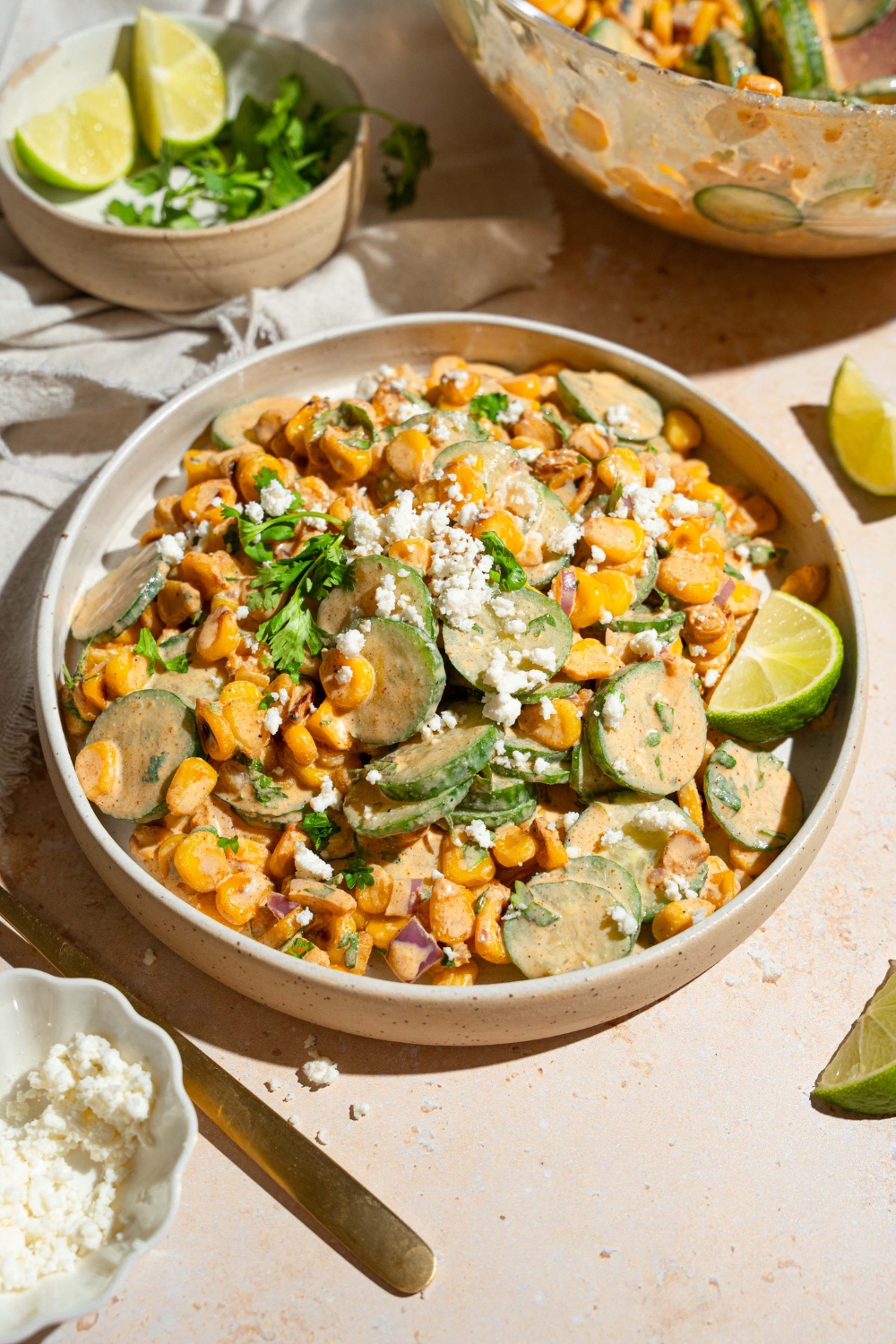 A white bowl with Mexican cucumber salad tossed with corn, cilantro, and onion tossed in an elote dressing and topped with cotija cheese. The bowl is on a tan counter with a bowl of cheese and bowl of toppings.