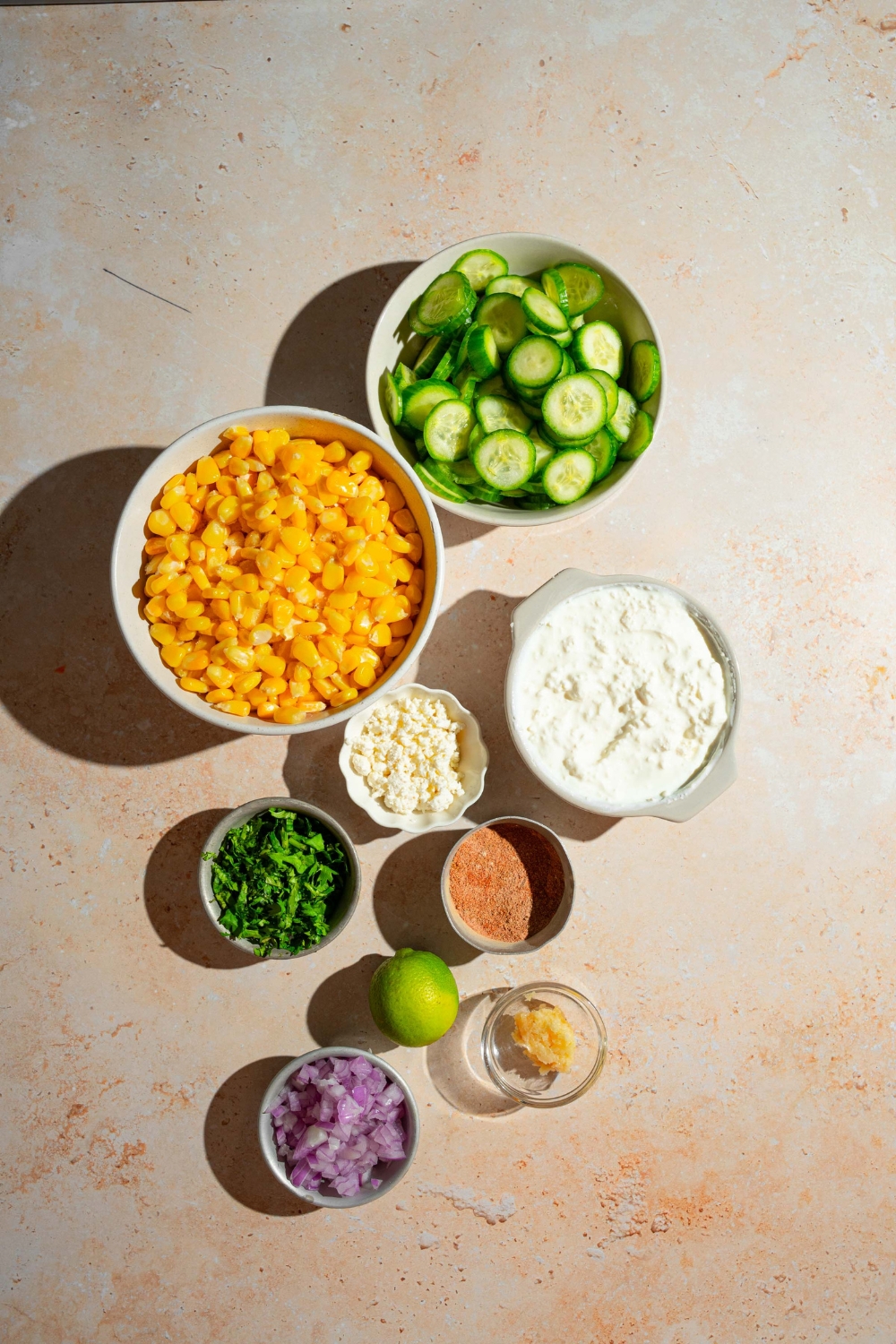 A tan counter with several bowls containing ingredients to make Mexican cucumber salad including corn, sliced cucumber, cottage cheese, cotija cheese, cilantro, red onion, elote seasoning, and more.