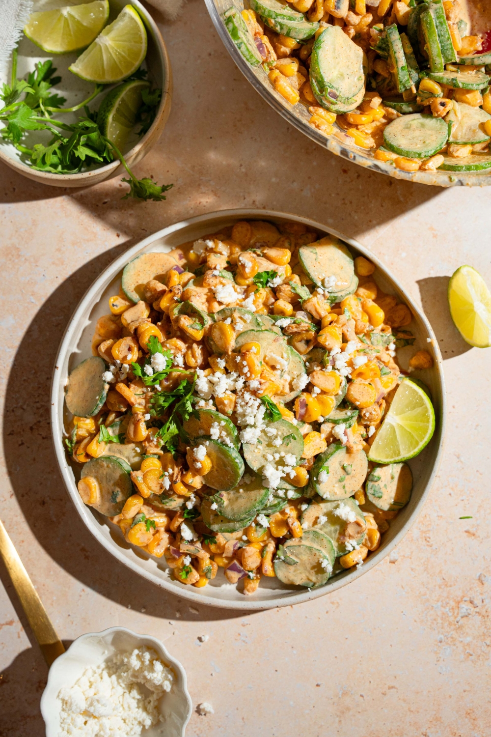 A white bowl with Mexican cucumber salad tossed with corn, cilantro, and onion tossed in an elote dressing and topped with cotija cheese. The bowl is on a tan counter with a bowl of cheese and a bowl of garnishes.