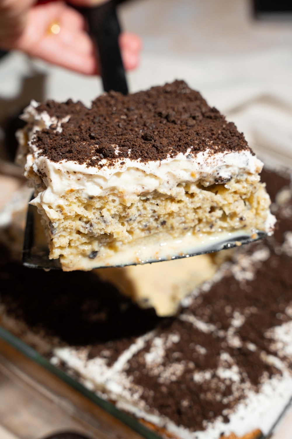 A close up of a spatula with a slice of Oreo tres leches cake. There is a dish of tres leches cake topped with whipped cream and crushed Oreos blurred in the background.
