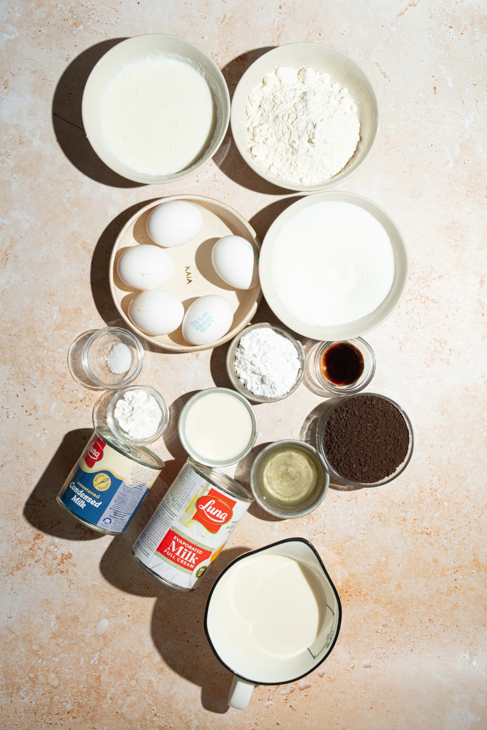 A tan counter with several bowls containing ingredients to make Oreo tres leches cake including crushed Oreos, sugar, milk, eggs, evaporated milk, condensed milk, vanilla, heavy whipping cream, and more.