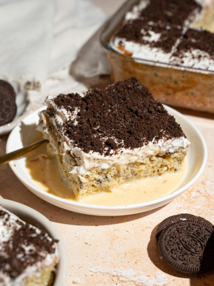 A white plate with a slice of Oreo tres leches cake on top of tres leches soak. There is a spoon on the plate. The plate is on a tan counter with Oreos and a baking dish with tres leches cake.