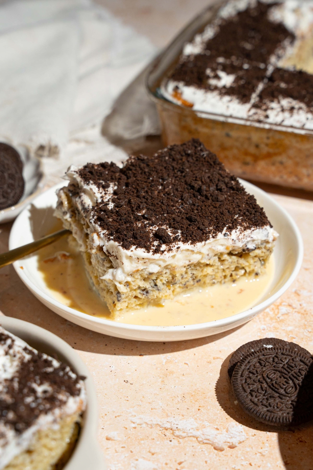A white plate with a slice of Oreo tres leches cake on top of tres leches soak. There is a spoon on the plate. The plate is on a tan counter with Oreos and a baking dish with tres leches cake.