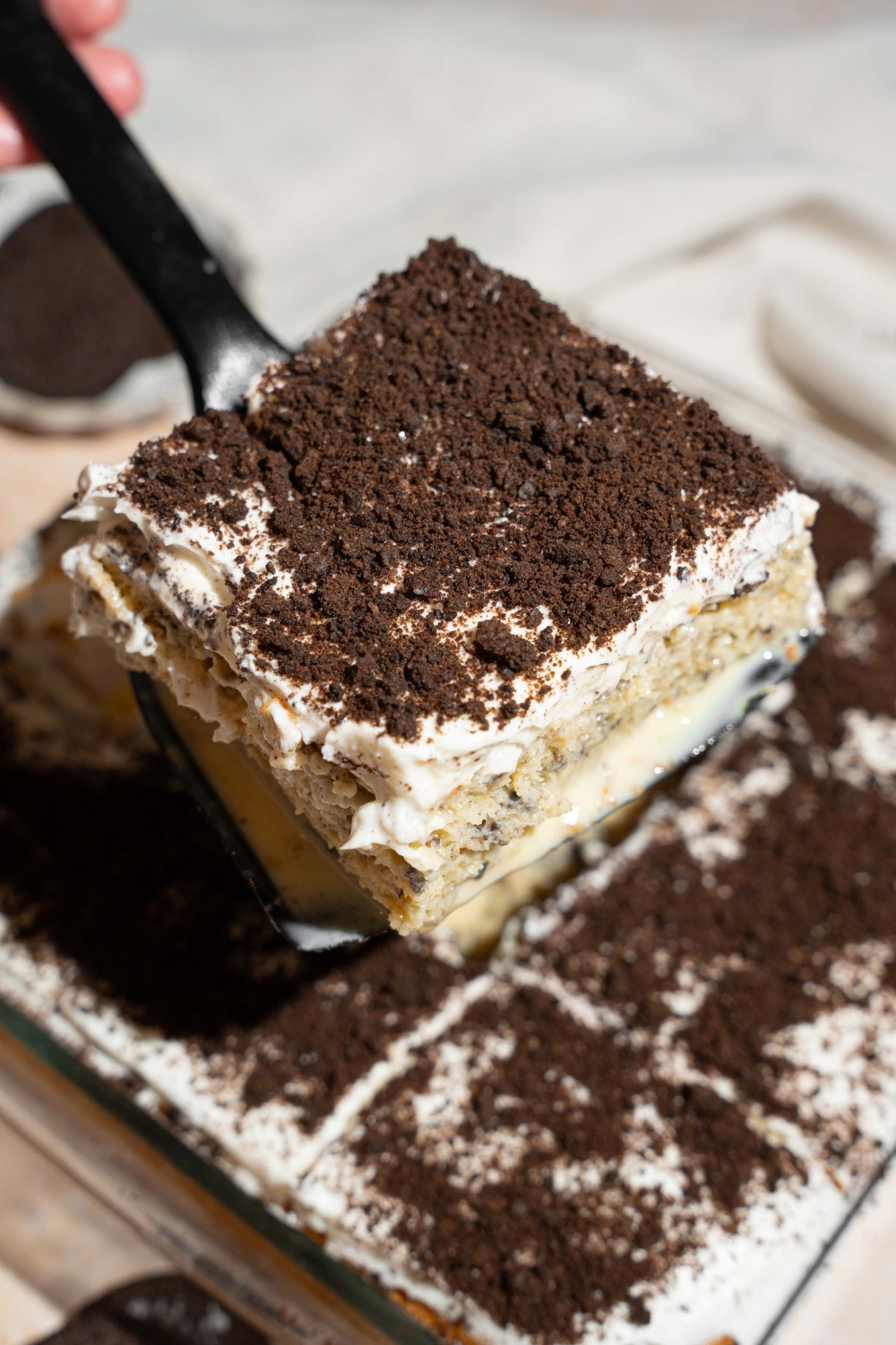 A close up of a spatula with a slice of Oreo tres leches cake. There is a dish of tres leches cake topped with whipped cream and crushed Oreos blurred in the background.