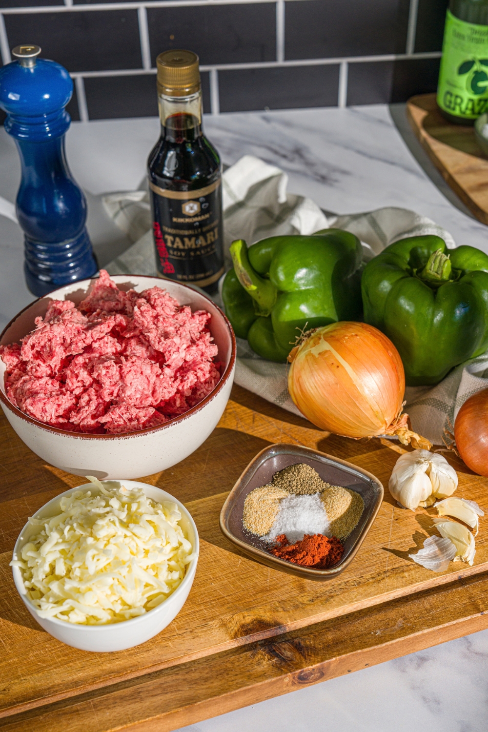 A wooden board with ingredients to make a philly cheesesteak bowl including ground beef, onion, green peppers, shredded cheese, garlic, and seasonings.