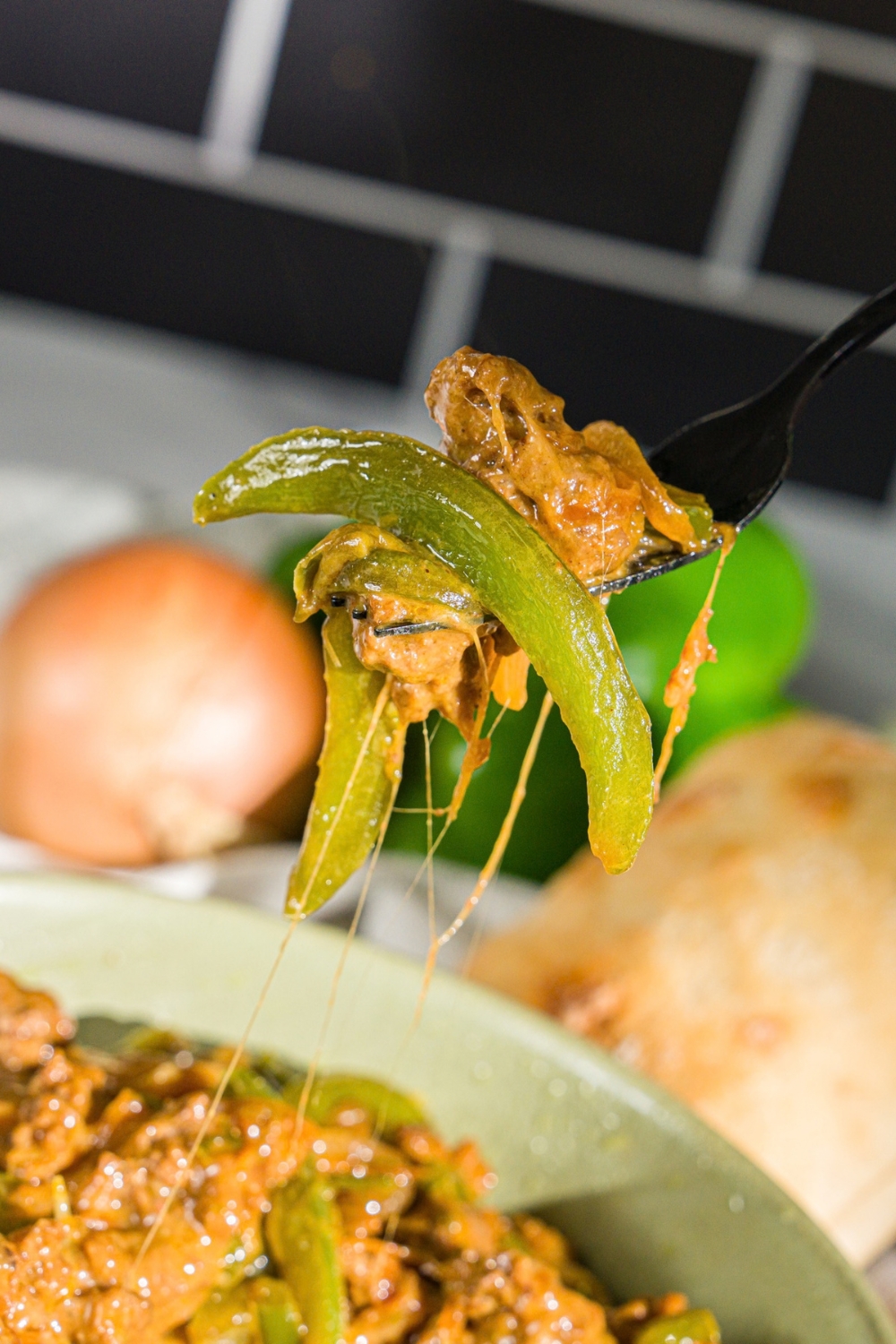 A close up of a fork with a bite from a philly cheesesteak bowl including ground beef, sliced green pepper, and cheese. There is a philly cheesesteak bowl blurred in the background.
