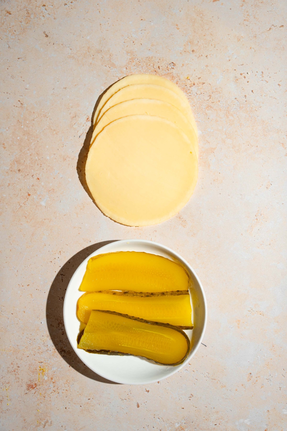 A tan counter with ingredients to make pickle bread including slices of cheese and a plate of sliced pickles.
