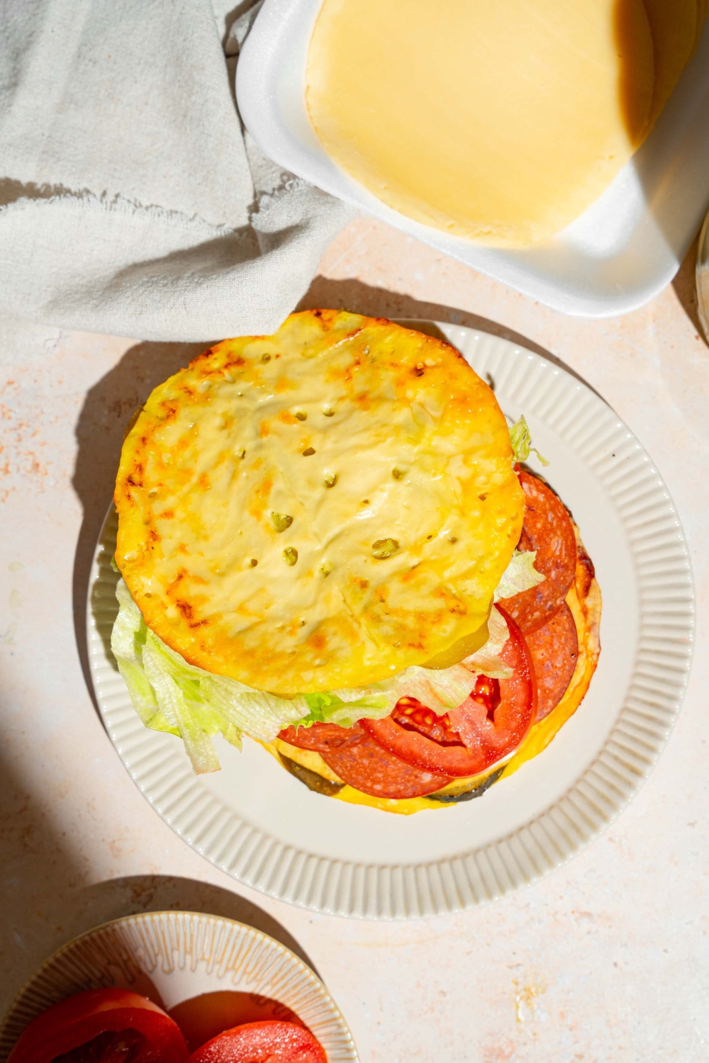 A white plate with a sandwich made with pickle bread, lettuce, tomato, and deli meat. The plate is on a tan counter with a white cloth napkin.