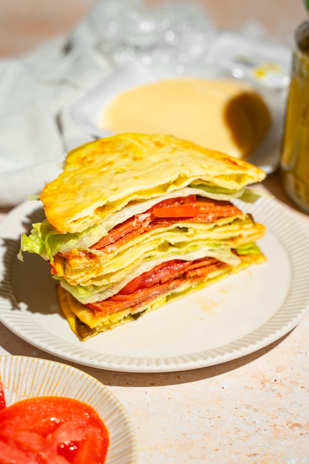 A white plate with a sandwich made with pickle bread, lettuce, tomato, and deli meat. The sandwich is sliced in half with one half stacked on the other. The plate is on a tan counter with a white cloth napkin.