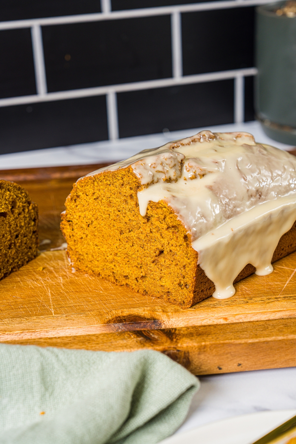 A wooden board with a loaf of pumpkin bread with maple glaze. The board is on a marble counter with a green cloth napkin.