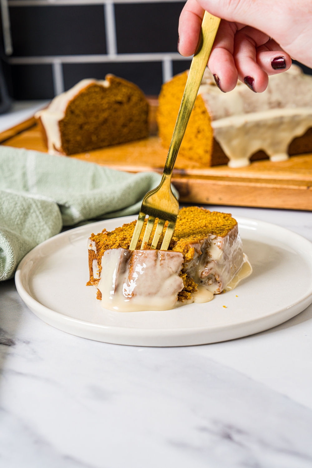 A white plate with a slice of pumpkin bread with maple glaze. There is a fork taking a bite from the slice. The plate is on a marble counter with a green cloth napkin.