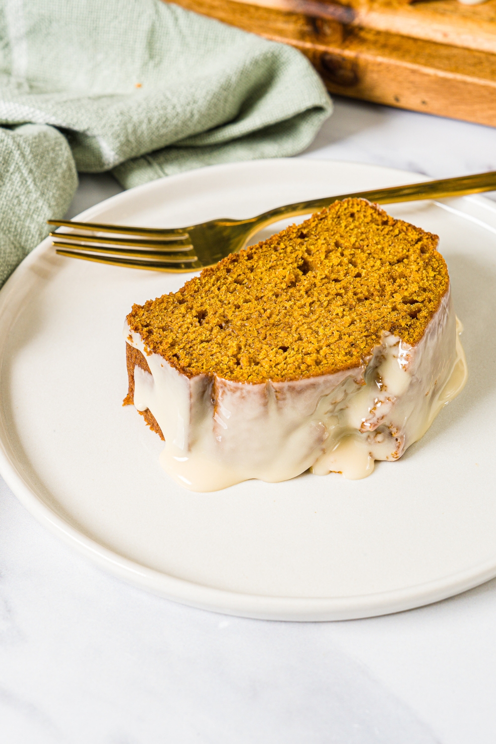 A white plate with a slice of pumpkin bread with maple glaze. There is a fork on the plate. The plate is on a marble counter with a green cloth napkin.