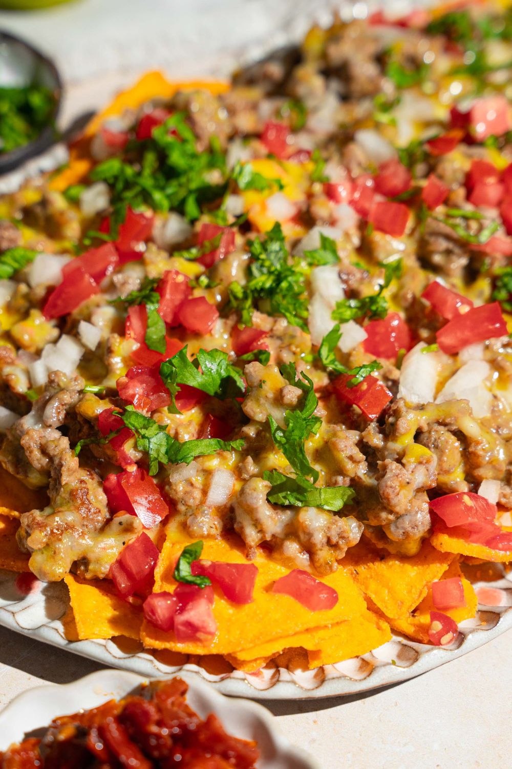 An oval platter with sliding sheet pan nachos topped with shredded cheese, ground beef, diced tomatoes and onions, and fresh parsley. The platter is on a tan counter.