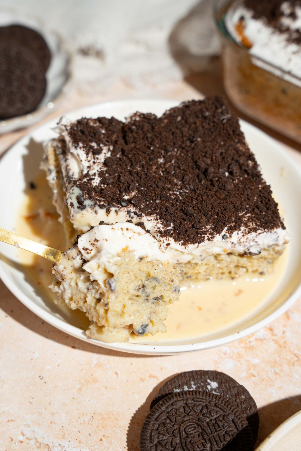 A white plate with a slice of Oreo tres leches cake on top of tres leches soak. A spoon is taking a bite from the slice. The plate is on a tan counter.