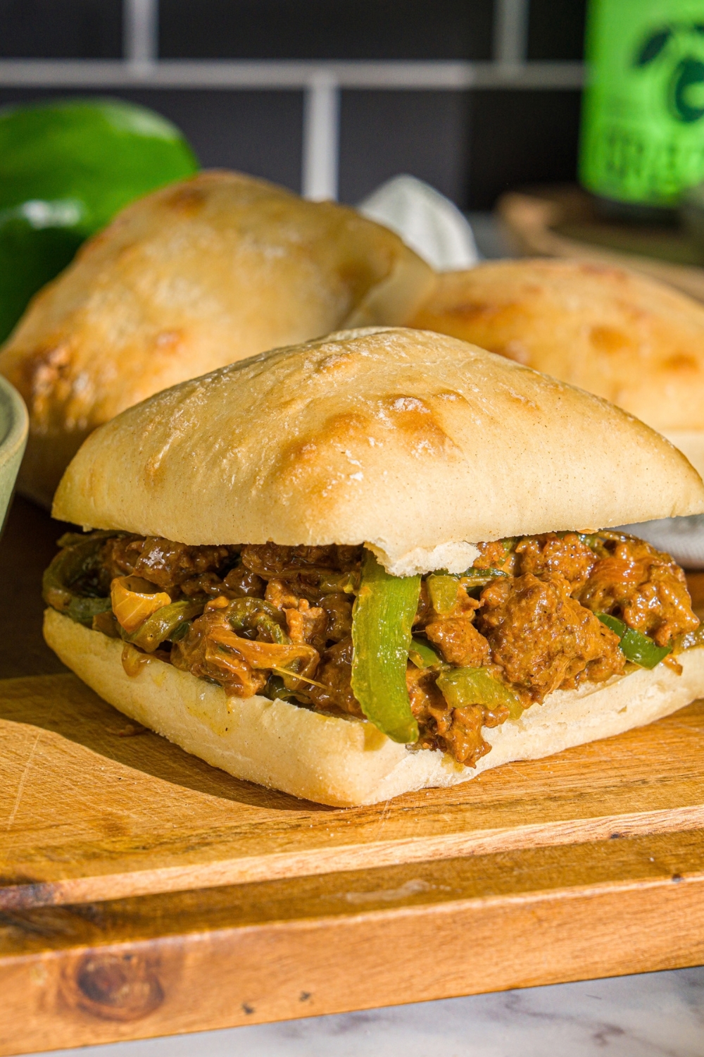A philly cheesesteak bowl mixture with ground beef, green peppers, and cheese on a ciabatta roll. The sandwich is on a wooden board with ciabatta rolls.