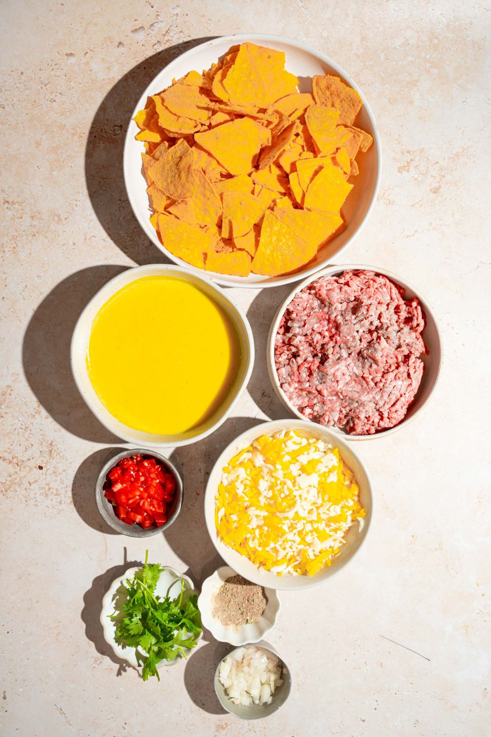 A tan counter with several bowls containing ingredients to make sliding sheet pan nachos including tortilla chips, nacho cheese, ground beef, shredded cheese, and more.