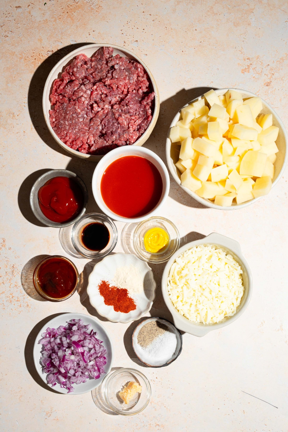 A tan counter with several bowls containing ingredients to make sloppy joe potato skillet including ground beef, potatoes, shredded cheese, onion, ketchup, mustard, garlic, and more.