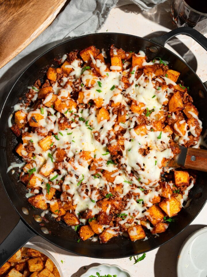 A skillet with sloppy joe potato skillet topped with melted cheese and garnished with fresh parsley. There is a spatula in the skillet. The skillet is on a tan counter with small bowls of ingredients.