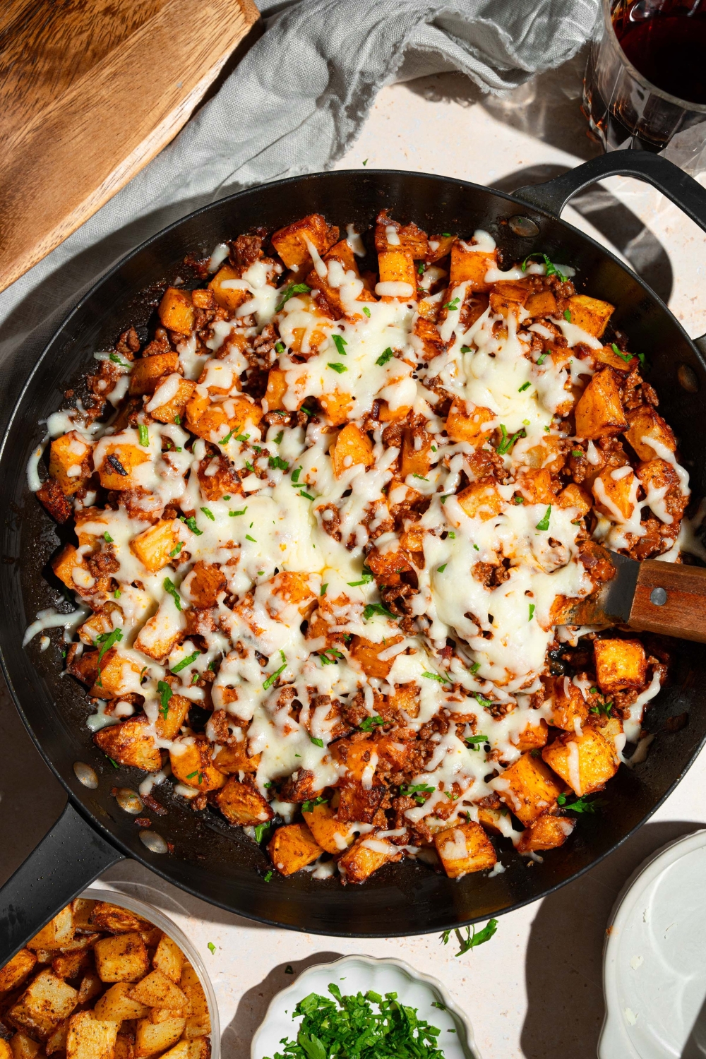 A skillet with sloppy joe potato skillet topped with melted cheese and garnished with fresh parsley. There is a spatula in the skillet. The skillet is on a tan counter with small bowls of ingredients.