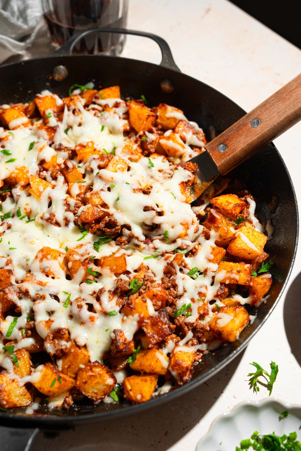 A skillet with sloppy joe potato skillet topped with melted cheese and garnished with fresh parsley. There is a spatula in the skillet. The skillet is on a tan counter.