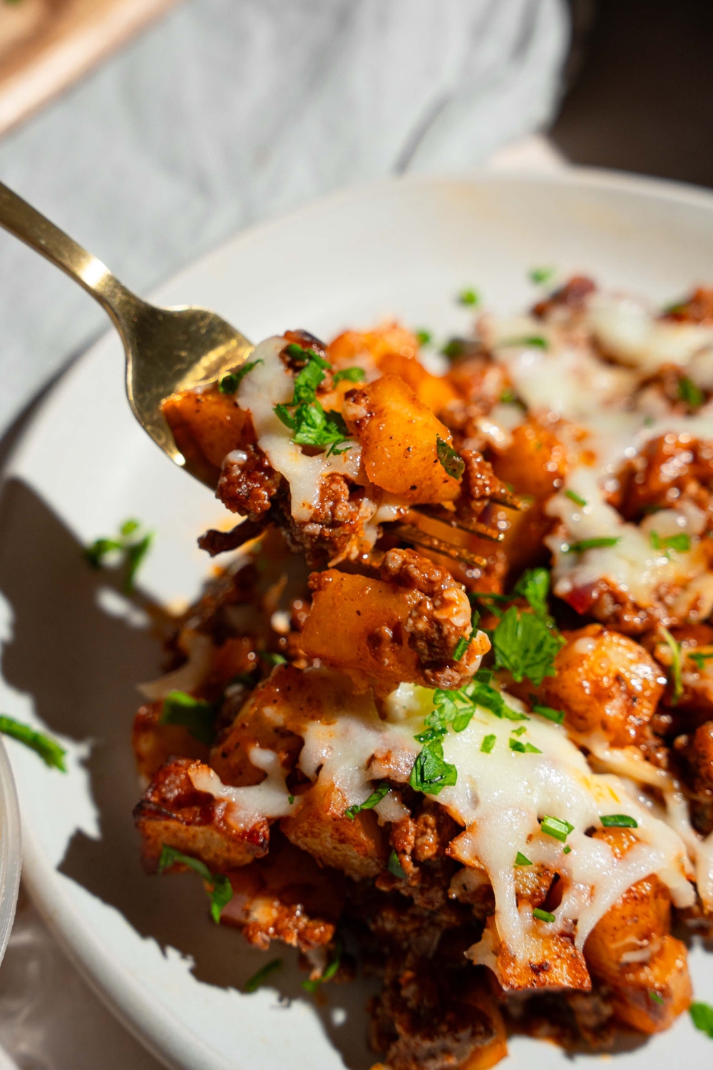 A white plate with sloppy joe potato skillet topped with melted cheese and garnished with fresh parsley. A fork is taking a bite from the bowl. The plate is on a tan counter.