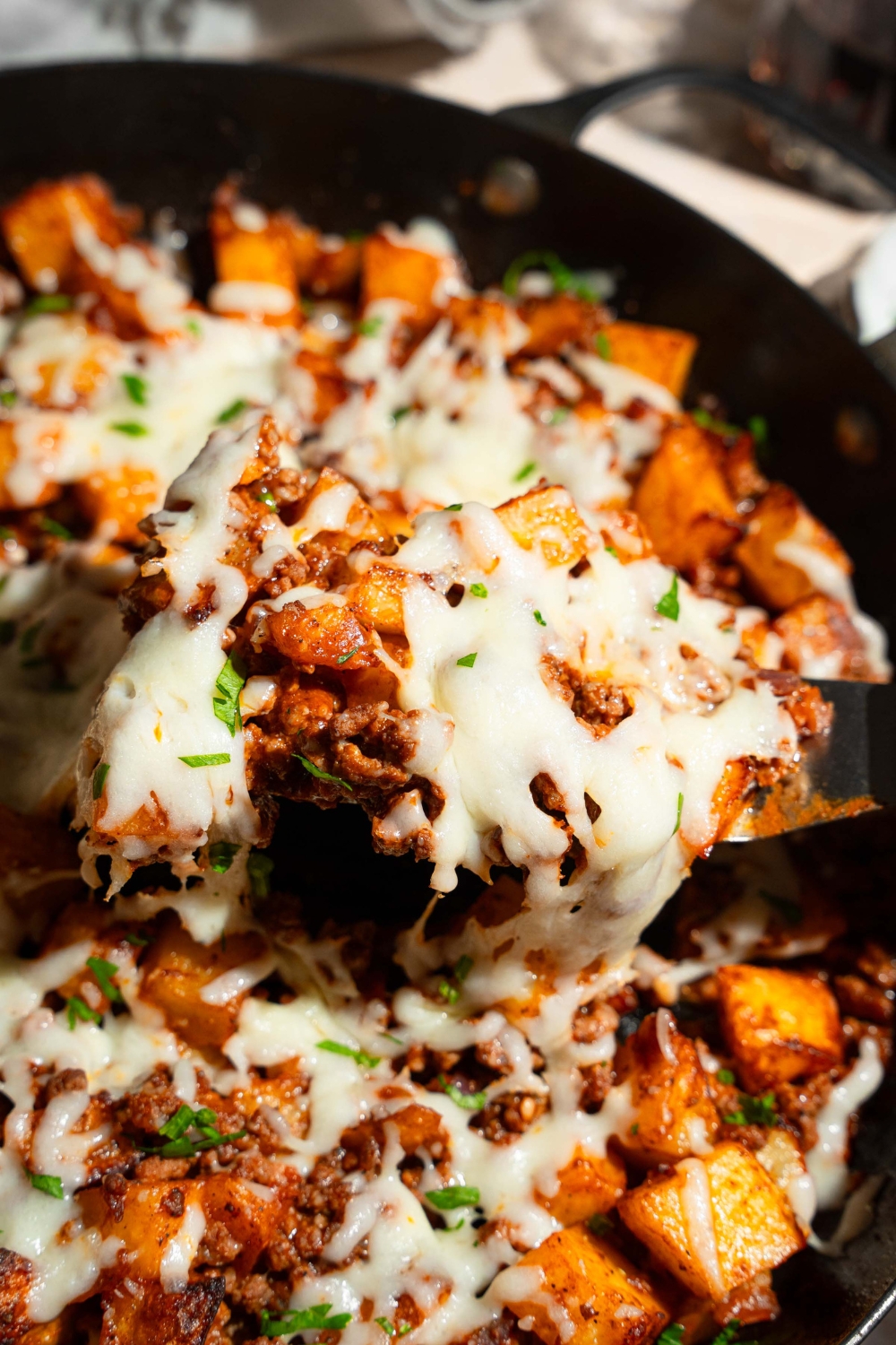A skillet with sloppy joe potato skillet topped with melted cheese and garnished with fresh parsley. There is a spatula in the skillet. The skillet is on a tan counter.
