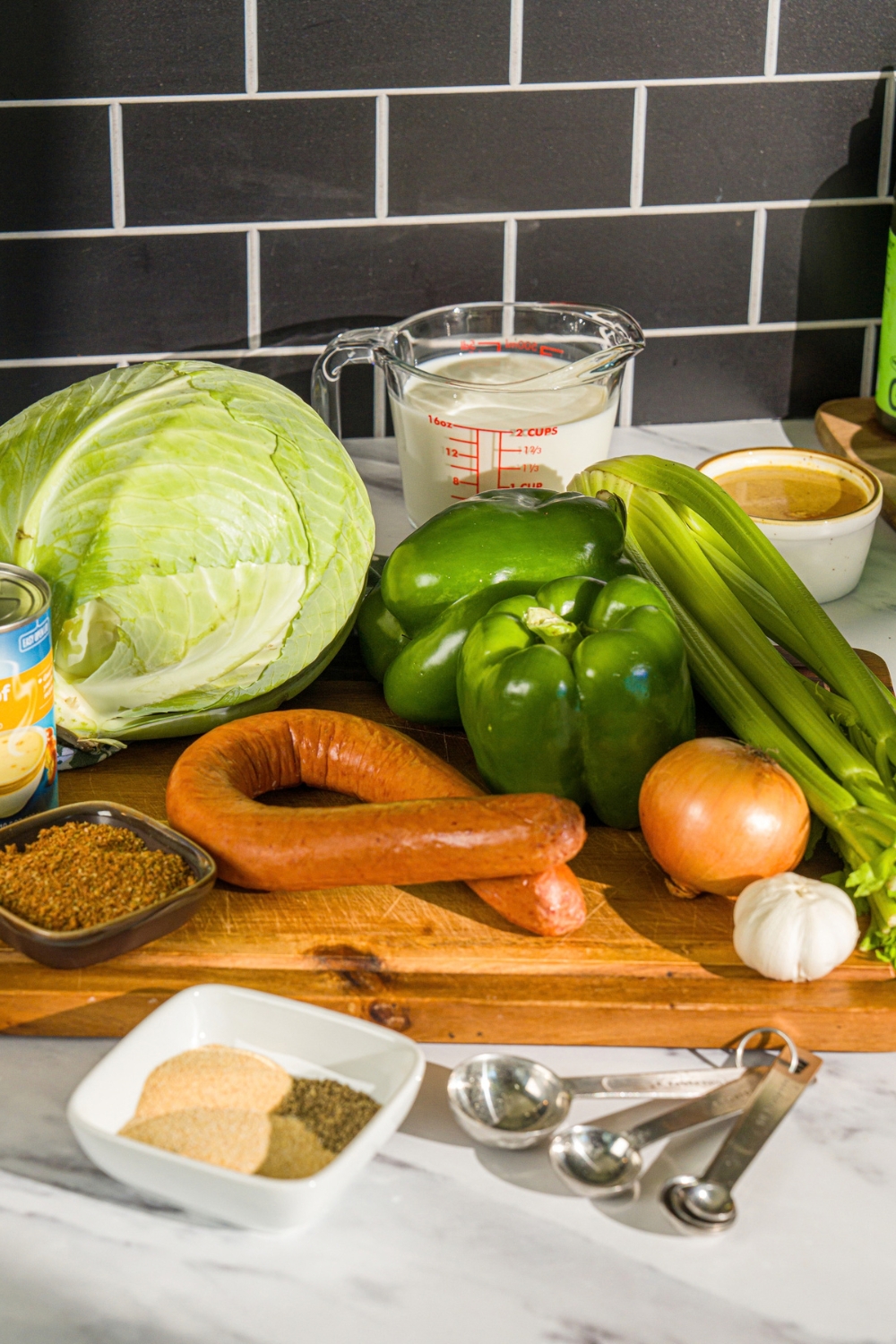 A wooden board with ingredients to make smothered cabbage including cabbage, green pepper, celery, onion, garlic, sausage, milk, and seasonings.