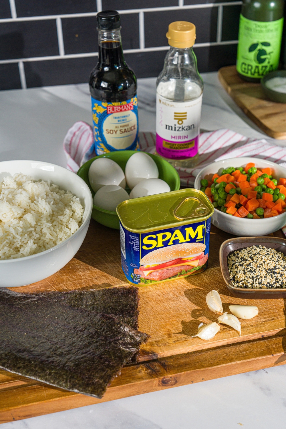 A wooden board with ingredients to make spam musubi bowl including spam, rice, garlic, peas and carrots, eggs, mirin, soy sauce, and seasonings.