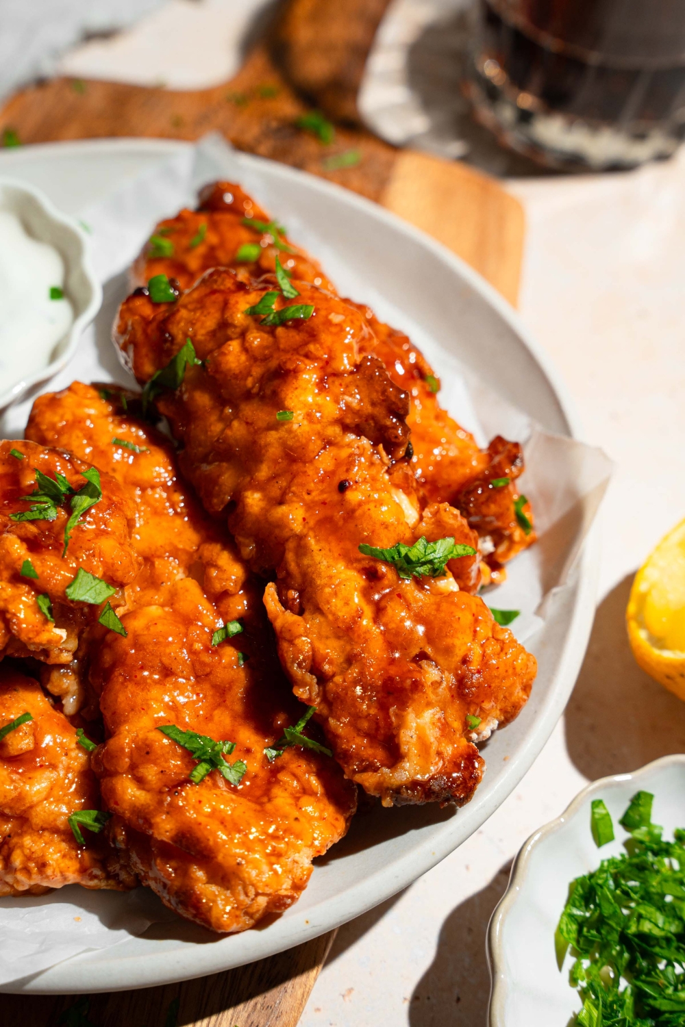 A white plate with sweet chili chicken tenders garnished with fresh parsley and served with a small bowl of dipping sauce. The plate is on a wooden board on a tan counter with garnishes.