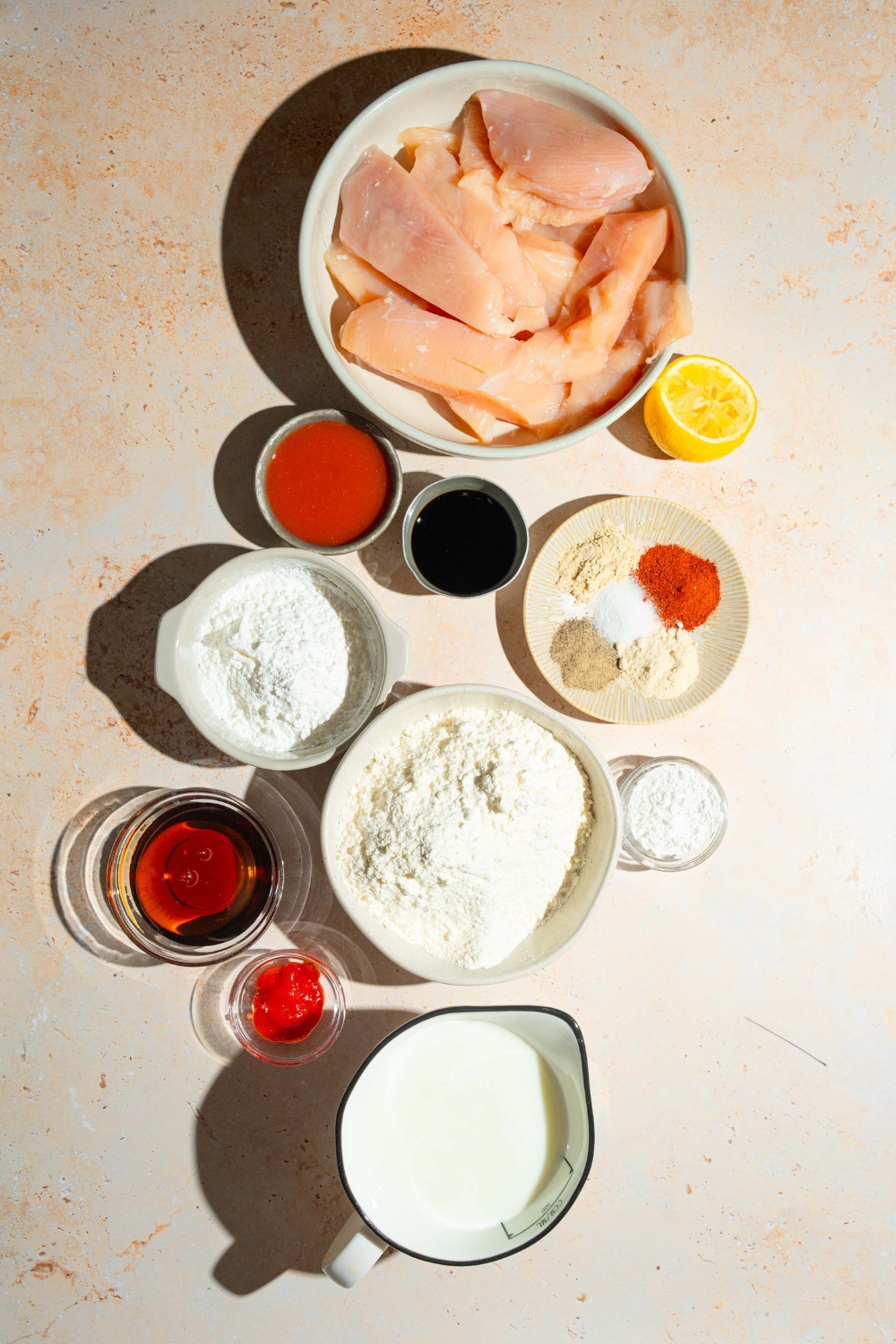 A tan counter with several bowls containing ingredients to make sweet chili chicken tenders including chicken tenders, buttermilk, hot sauce, soy sauce, lemon, flour, honey, ketchup, chili garlic sauce, and more.