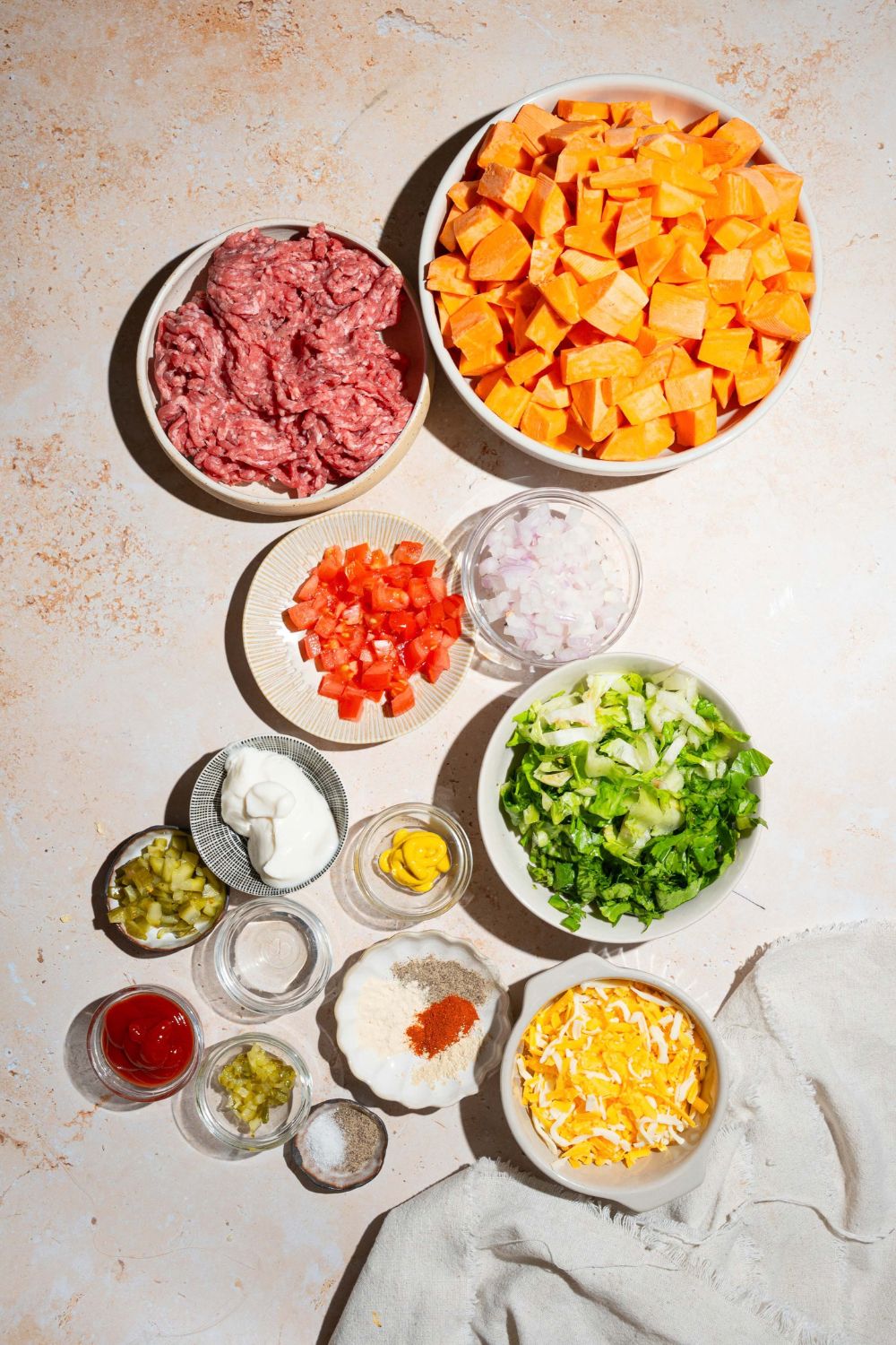 A tan counter with several bowls containing ingredients to make sweet potato burger bowls including ground beef, sweet potatoes, tomatoes, shredded lettuce, shredded cheese, onion, relish, mayo, ketchup, and more.