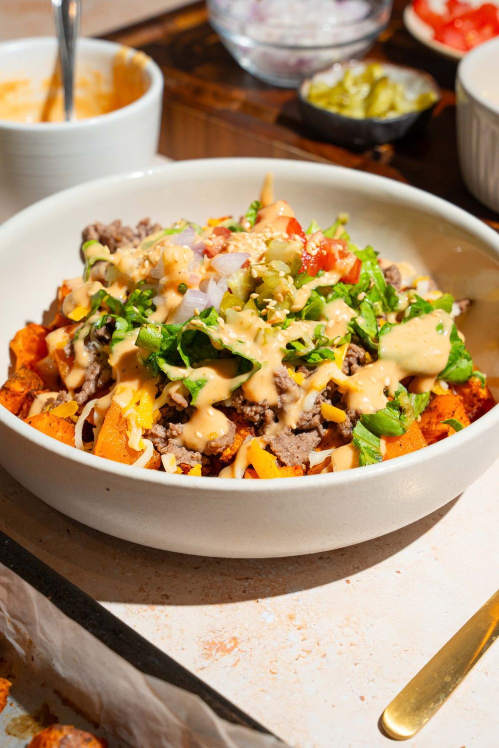 A white bowl with a sweet potato burger bowl including sweet potatoes, ground beef, shredded lettuce, shredded cheese, and tomatoes drizzled with burger sauce The bowl is on a tan counter with bowls of ingredients.