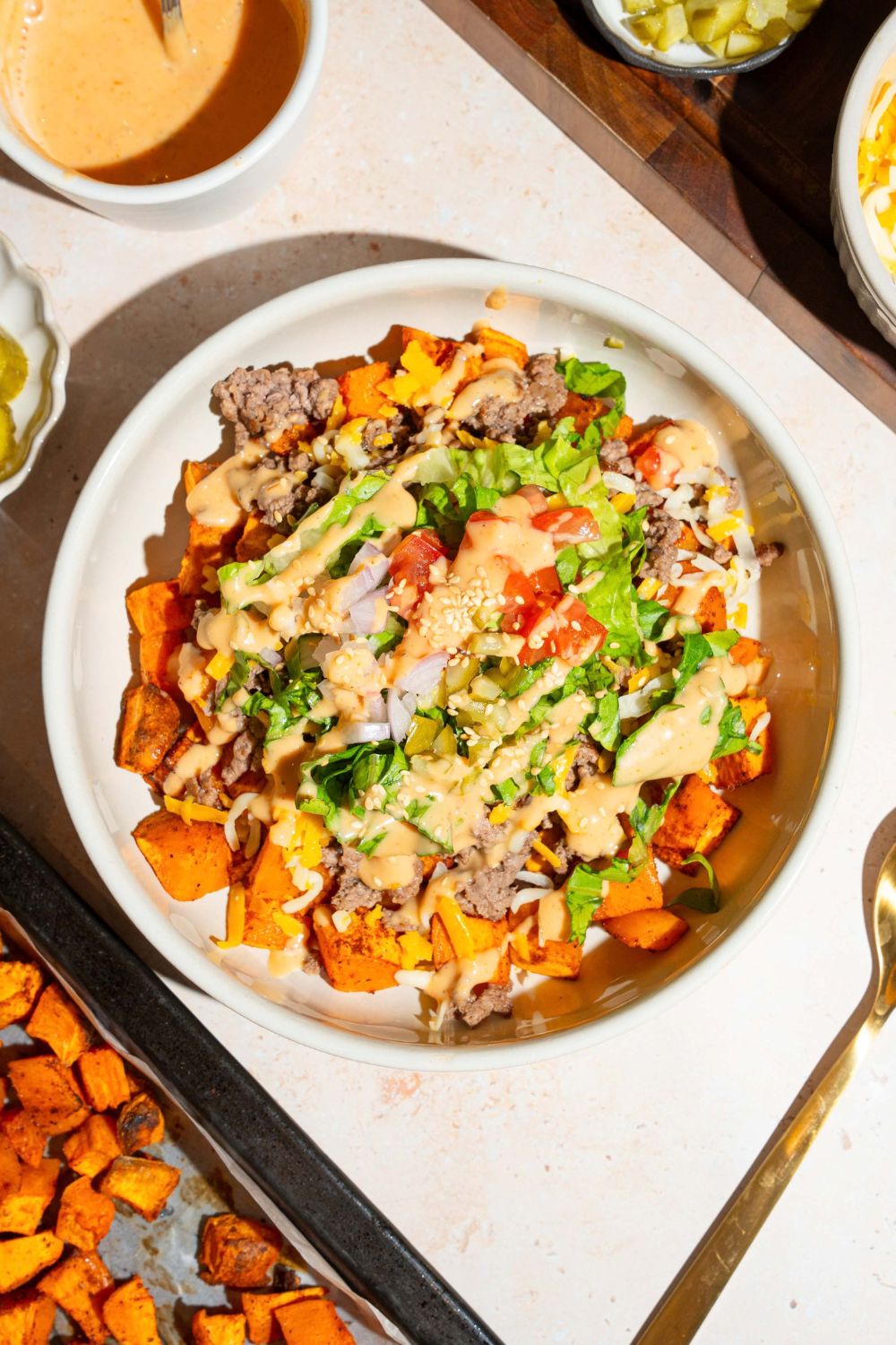 A white bowl with a sweet potato burger bowl including sweet potatoes, ground beef, shredded lettuce, shredded cheese, and tomatoes drizzled with burger sauce The bowl is on a tan counter with bowls of ingredients.