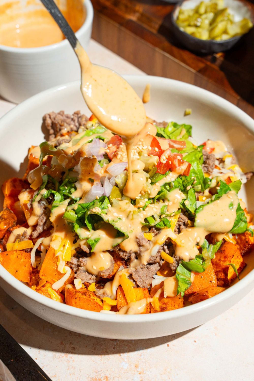 A white bowl with a sweet potato burger bowl including sweet potatoes, ground beef, shredded lettuce, shredded cheese, and tomatoes. A spoon is drizzling burger sauce over the bowl. The bowl is on a tan counter with bowls of ingredients.