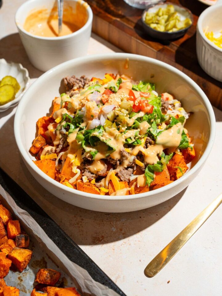 A white bowl with a sweet potato burger bowl including sweet potatoes, ground beef, shredded lettuce, shredded cheese, and tomatoes drizzled with burger sauce The bowl is on a tan counter with bowls of ingredients.