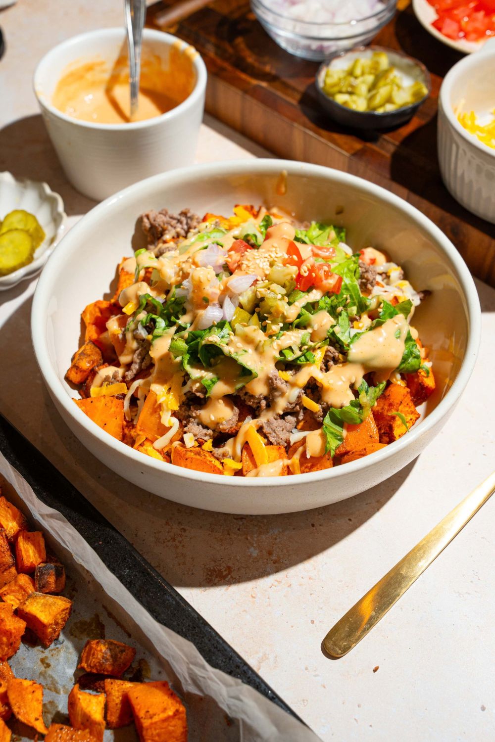 A white bowl with a sweet potato burger bowl including sweet potatoes, ground beef, shredded lettuce, shredded cheese, and tomatoes drizzled with burger sauce The bowl is on a tan counter with bowls of ingredients.