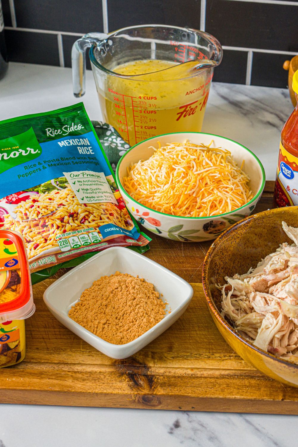 A wooden board with ingredients to make swim queso chicken and rice including Knorr Mexican rice packets, shredded chicken, queso cheese, shredded cheese, and more.