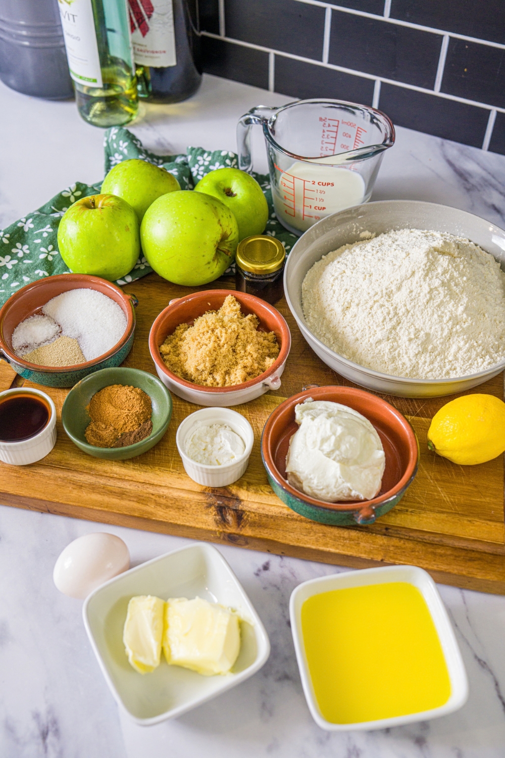 A wooden board with ingredients to make twisted apple pie rolls including flour, eggs, yogurt, butter, lemon, apples, brown sugar, cinnamon, and more.