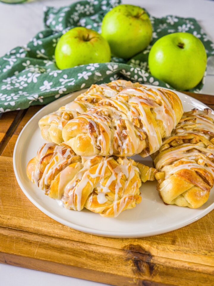 A white plate with twisted apple pie rolls drizzled with icing. The plate is on a wooden board with a green cloth napkin and green apples.