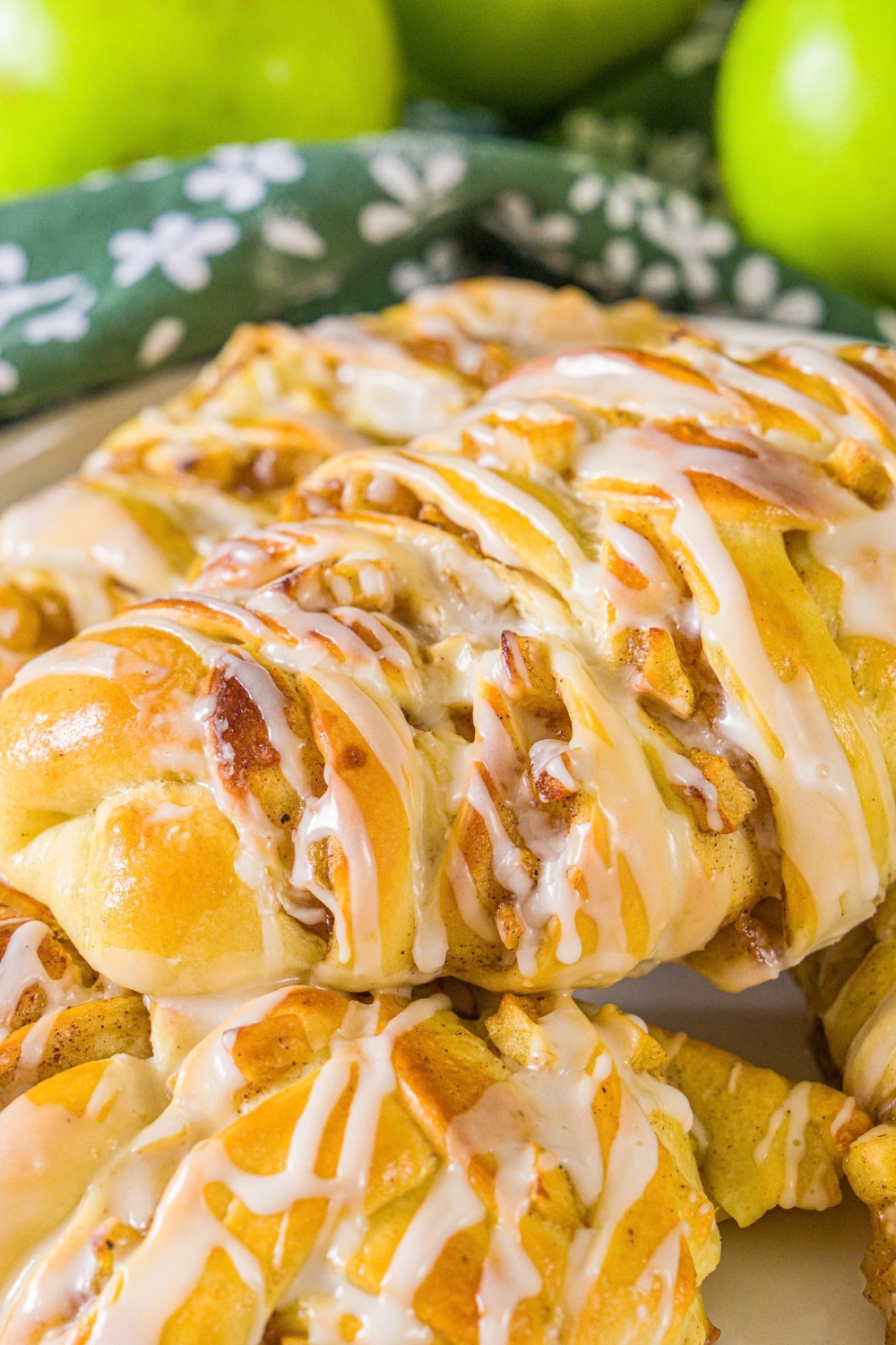 A white plate with twisted apple pie rolls drizzled with icing. The plate is on a wooden board with a green cloth napkin and green apples.
