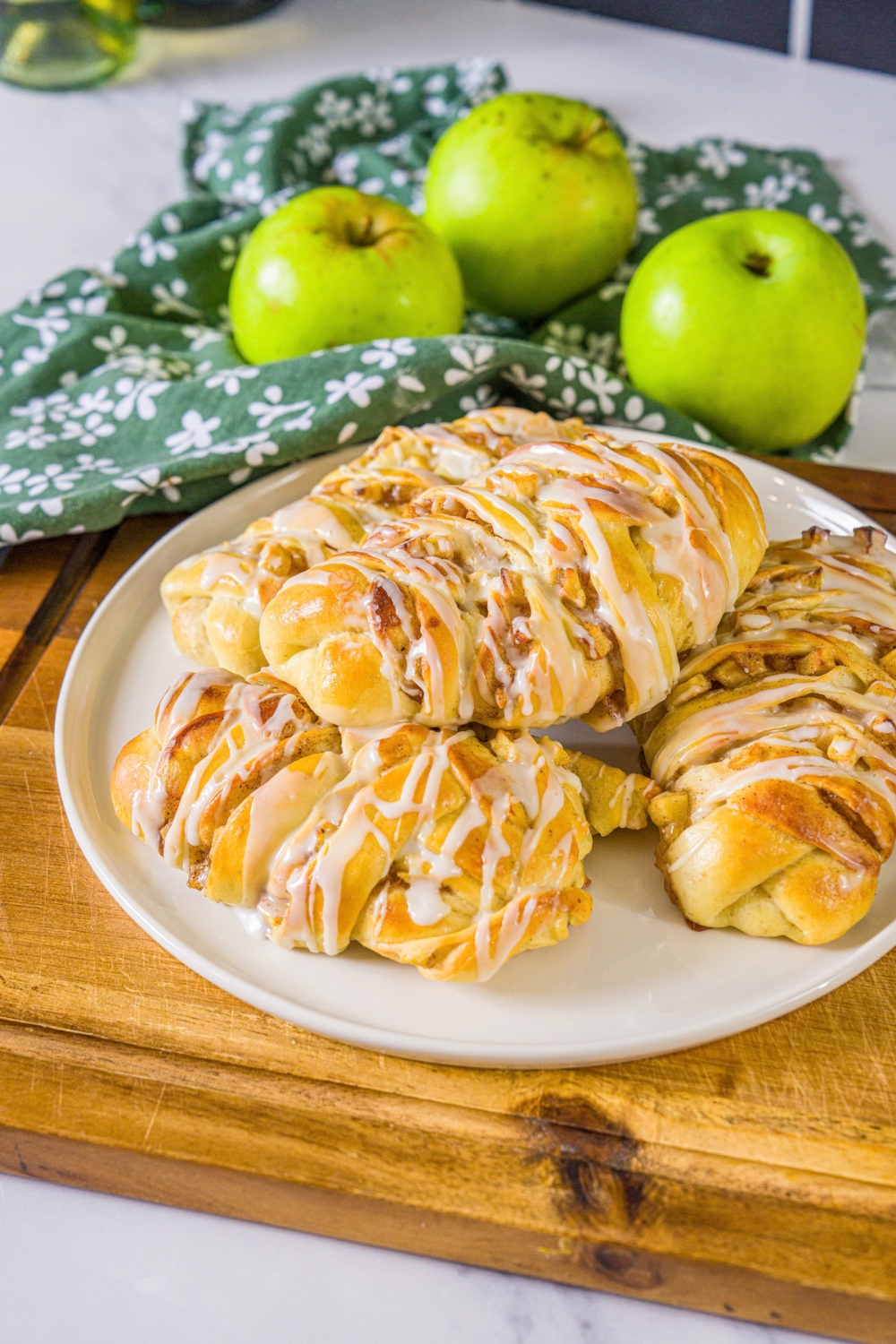 A white plate with twisted apple pie rolls drizzled with icing. The plate is on a wooden board with a green cloth napkin and green apples.