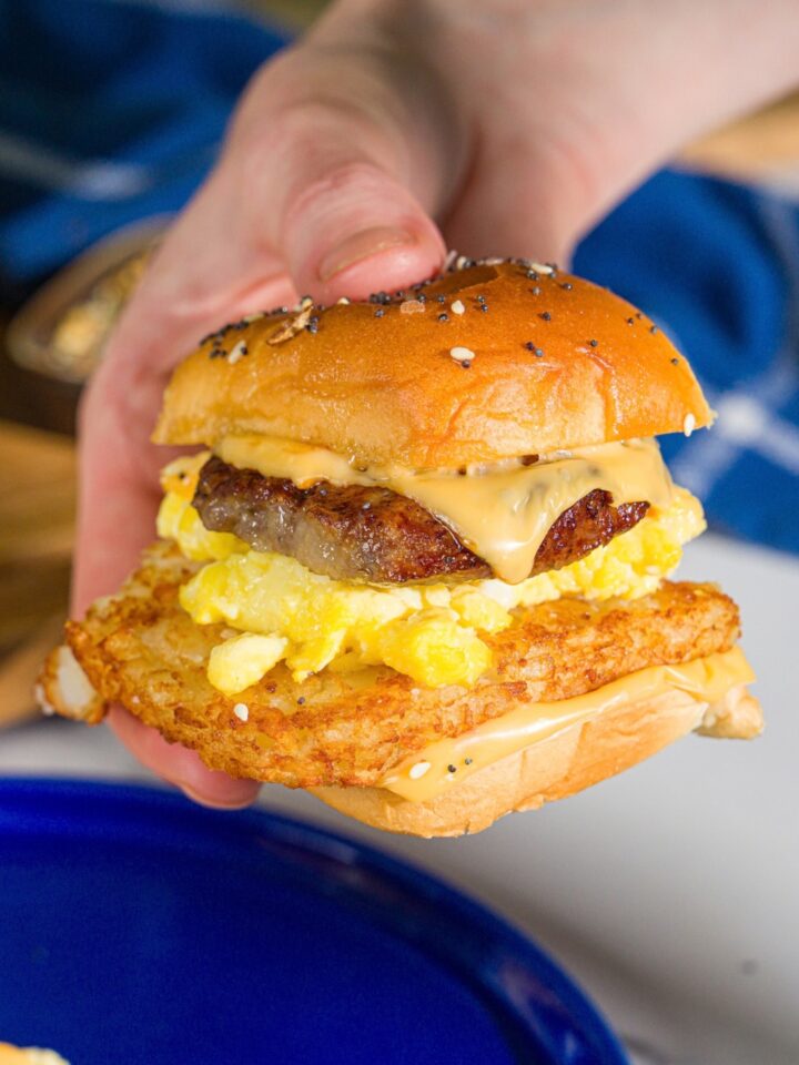 A hand holding a breakfast slider made with hashbrowns, scrambled eggs, sausage, and cheese on a seasoned hawaiian roll.
