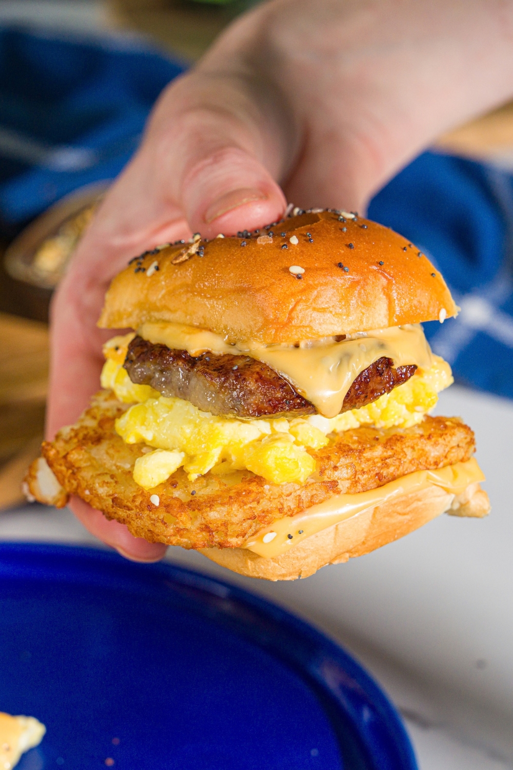 A hand holding a breakfast slider made with hashbrowns, scrambled eggs, sausage, and cheese on a seasoned hawaiian roll.