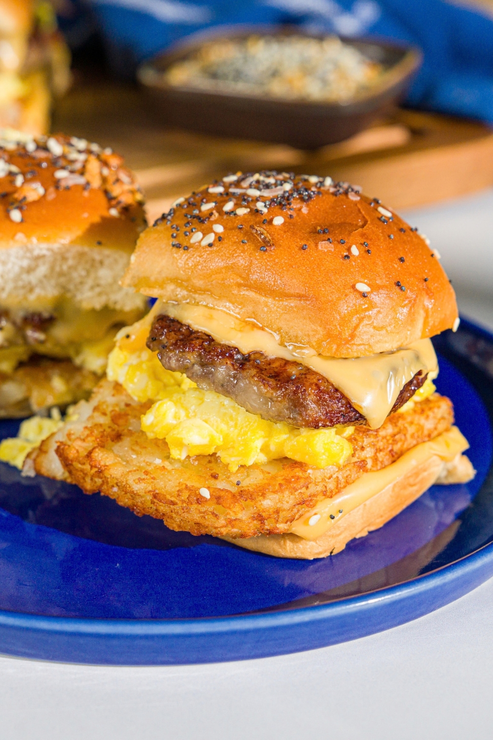 A blue plate with two breakfast sliders made with sausage, hashbrowns, scrambled eggs, and cheese on a seasoned hawaiian roll. The plate is on a marble counter.