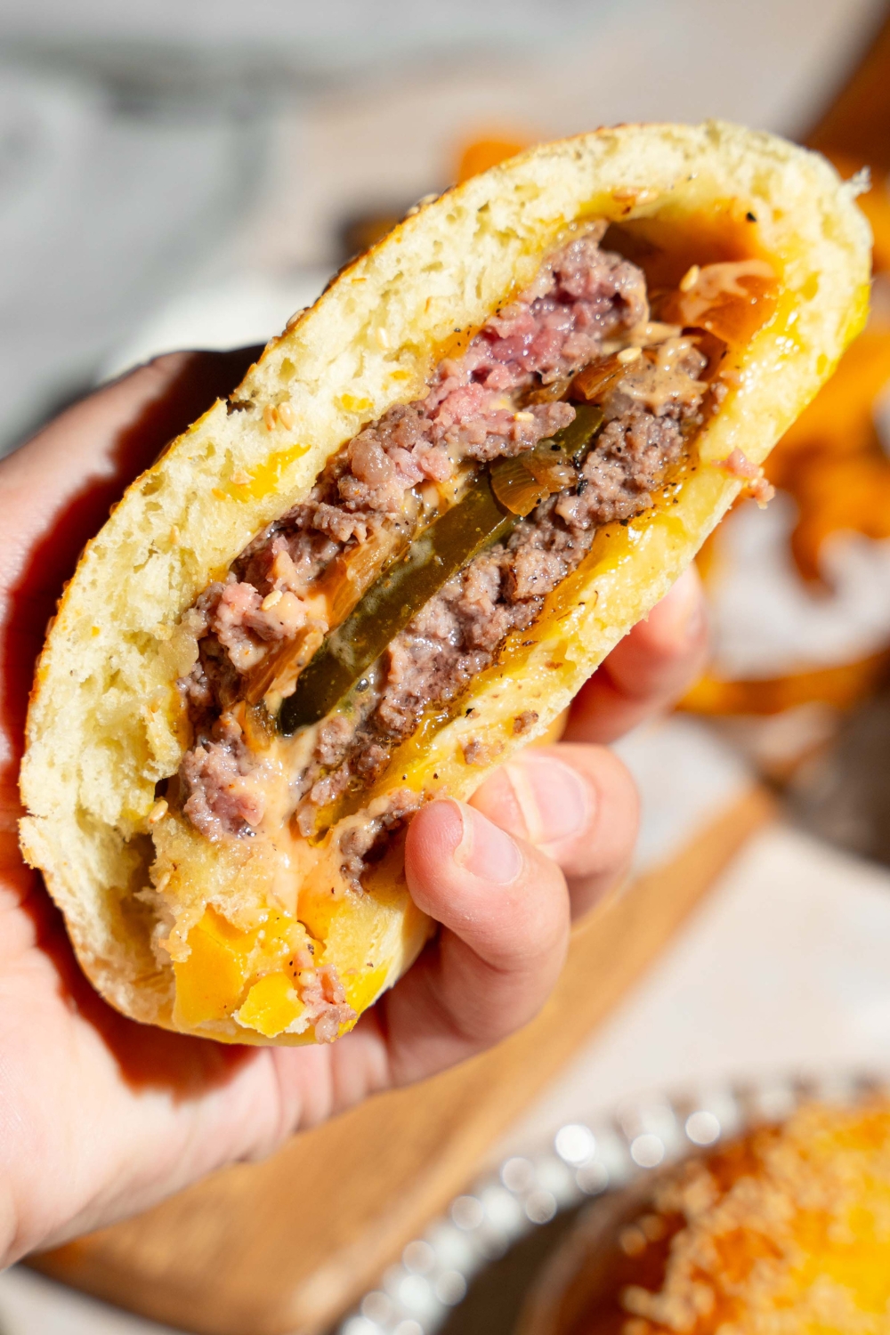 A close up of a hand holding a burger stuffed bun sliced in half. There is a wooden board on a tan counter blurred in the background.
