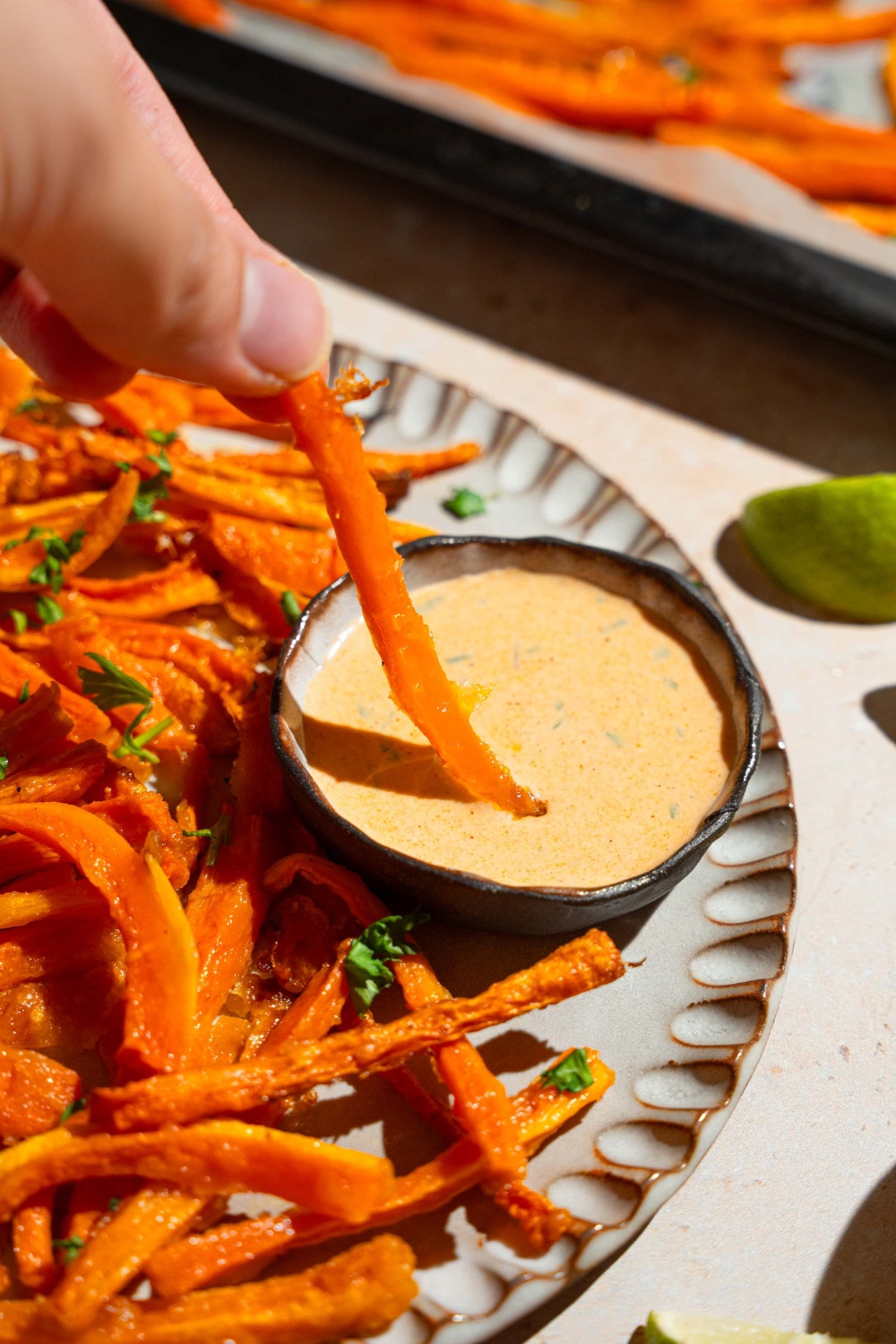 A plate of carrot fries garnished with fresh parsley and served with a bowl fo dipping sauce. A fry is being dipped in the sauce. The plate is on a tan counter with a baking sheet of fries and a white cloth napkin.