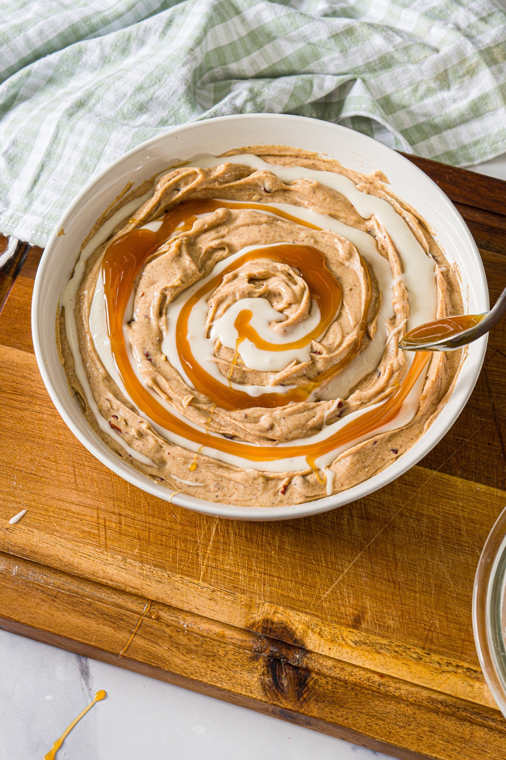 A spoon drizzling caramel sauce on top of cinnamon roll dip in a white bowl.
